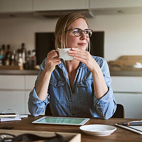 Frau sitzt mit Kaffee-Tasse am Schreibtisch und schaut in die Ferne